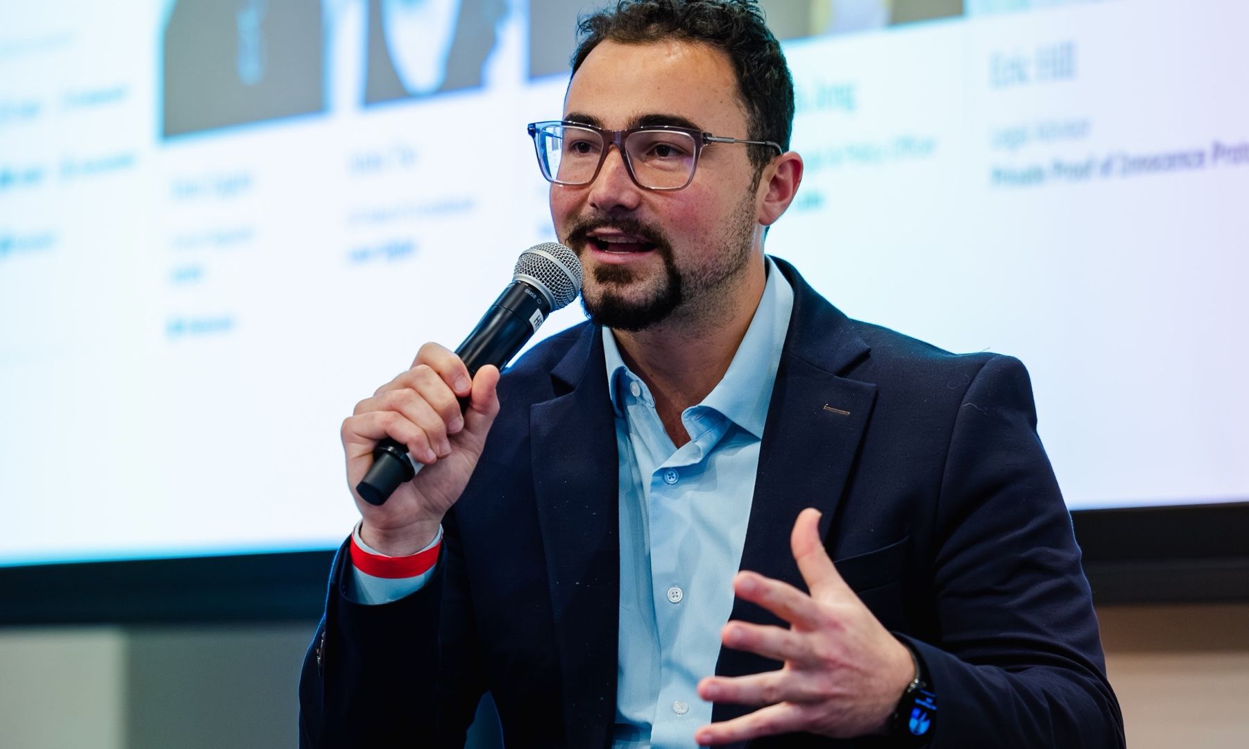 Dominic moderating the panel, holding a microphone and gesturing, with the speaker slide behind him