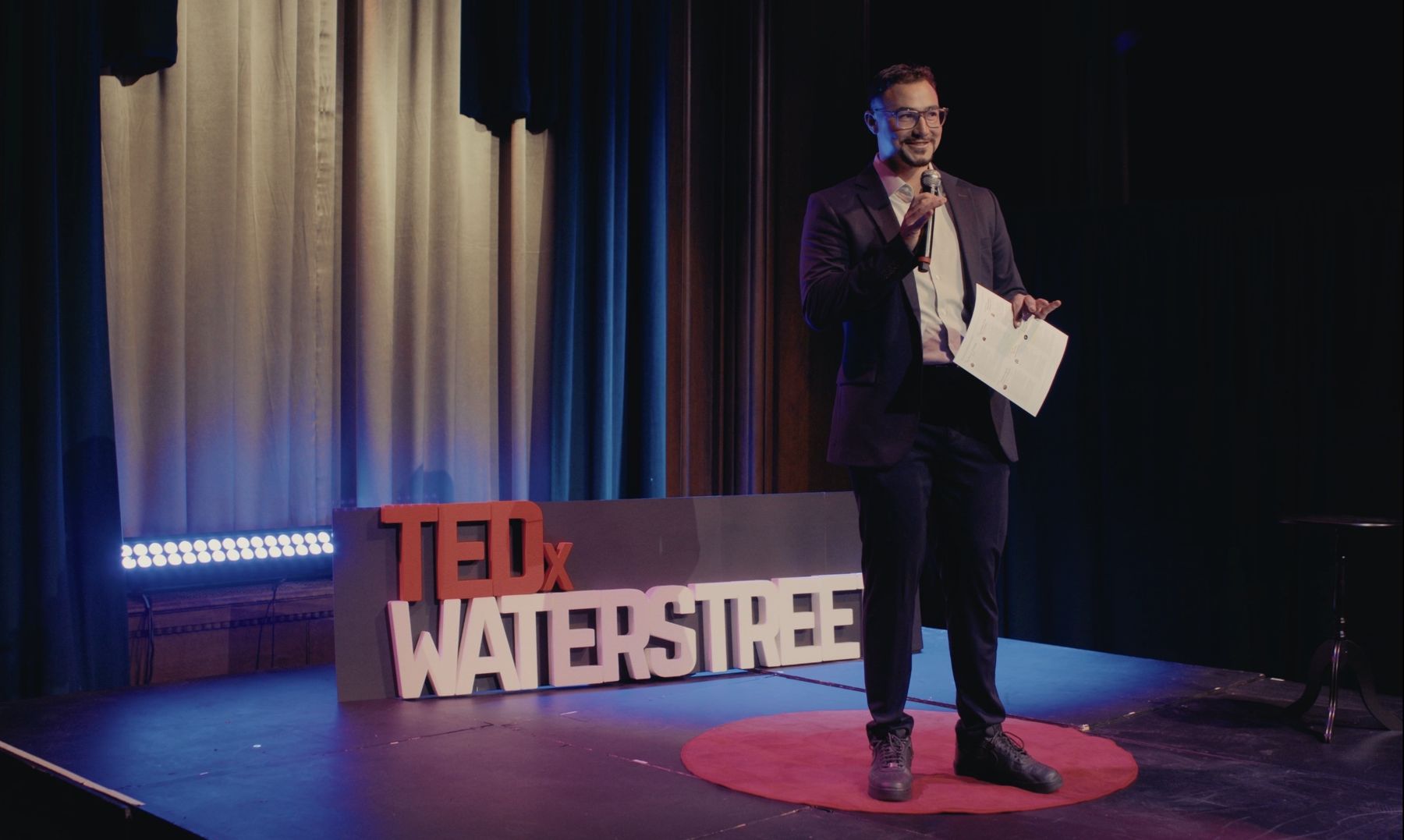Dominic hosting TEDxWaterStreet on stage beside the TEDxWaterStreet sign and red circular carpet